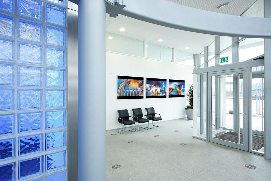 Ruhiger Empfangsbereich mit Glaswand und mehreren Sitzplätzen. An der Wand hängen drei Bilder mit lebhaften Farben. Die Eingangstür ist aus Glas, und in der Ecke steht eine Pflanze.