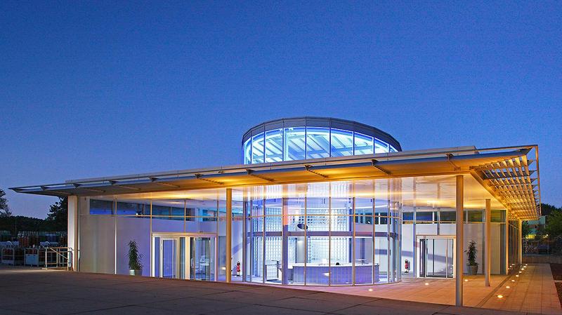 Moderne Architektur eines Gebäudes bei Dämmerung. Die Fassade ist aus Glas und Metall, mit einem runden Dach. Sanfte Beleuchtung hebt die Struktur hervor und schafft eine einladende Atmosphäre. Um das Gebäude herum sind Pflanzen und eine gepflasterte Fläche sichtbar.