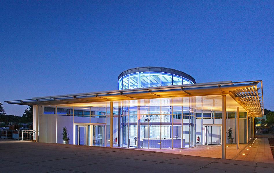 Moderne Architektur eines Gebäudes bei Dämmerung. Die Fassade ist aus Glas und Metall, mit einem runden Dach. Sanfte Beleuchtung hebt die Struktur hervor und schafft eine einladende Atmosphäre. Um das Gebäude herum sind Pflanzen und eine gepflasterte Fläche sichtbar.