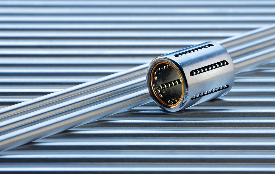 Ein Metallrohr liegt horizontal auf einer gestreiften Unterlage, während ein runder Metallzylinder darüber positioniert ist. Der Zylinder hat seitliche Öffnungen und einen glänzenden Korpus, was auf mechanische Funktionen hinweist.