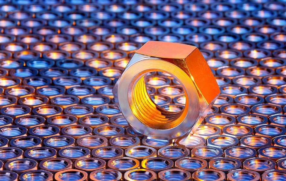 Eine große, glänzende Metallmutter steht auf einer Oberfläche aus vielen kleinen, runden metallischen Elementen. Das Licht reflektiert sich von der Mutter und den darunterliegenden Teilen, was zu einem schönen Farbspiel führt.