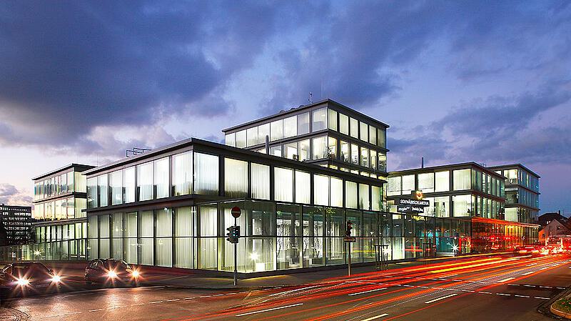 Moderne Bürogebäude mit Glasfassade bei Nacht, beleuchtet von Innen. Straßenverkehr ist sichtbar, mit Lichtspuren von fahrenden Autos. Der Himmel ist bewölkt und es ist Dämmerung.