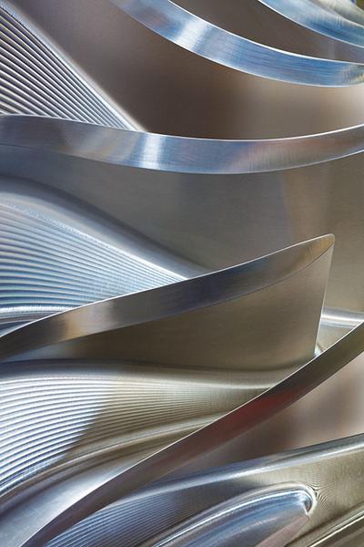 Eine Nahaufnahme von geschwungenen, polierten Metalloberflächen. Die Struktur zeigt wellenförmige und gebogene Elemente, die miteinander verbunden sind und verschiedene Lichtreflexionen erzeugen. Das Bild vermittelt eine dynamische und moderne Ästhetik durch die Kombination von Linien und Form.