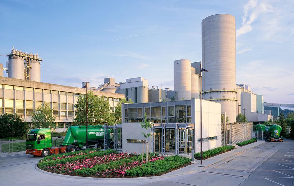 Ein modernes Industriegebäude mit großen Silos und einem Empfangsgebäude im Vordergrund. Grüne Lastwagen stehen vor dem Gebäude, umgeben von gepflegten Blumen und Bäumen. Der Himmel ist blau mit einigen Wolken.