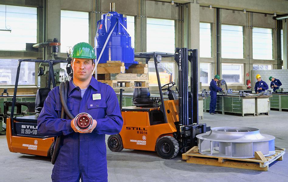 Ein Arbeiter in blauer Arbeitskleidung und mit einem grünen Helm steht in einer Industriehalle. Er hält ein Werkzeug in der Hand. Im Hintergrund sind Gabelstapler und weitere Arbeiter zu sehen, die an Maschinen arbeiten.