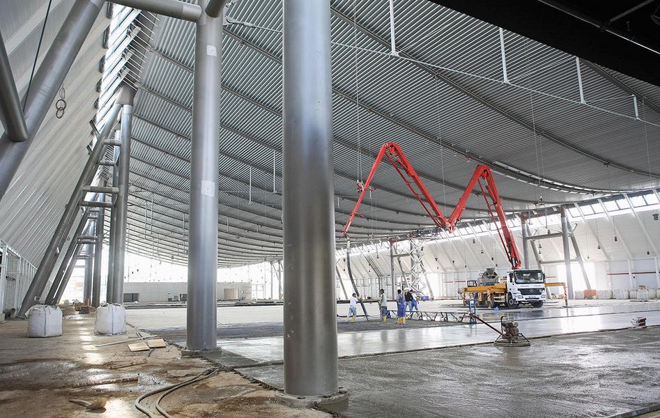 Ein großer, leerer Innenraum mit hohen, metallic schimmernden Wänden und einem gewölbten Dach. In der Mitte befindet sich ein Betonmischer mit einem langen Schlauch, während Arbeiter auf dem Boden beschäftigt sind. Stützen aus Edelstahl sind sichtbar.