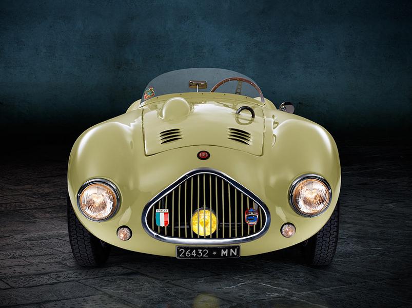 Ein klassisches, gelbgrünes Sportauto mit runder Form und zwei schmalen Scheinwerfern steht auf einem grauen, gepflasterten Boden. Im Hintergrund ist eine dunkle Wand sichtbar, die eine elegante Atmosphäre schafft.