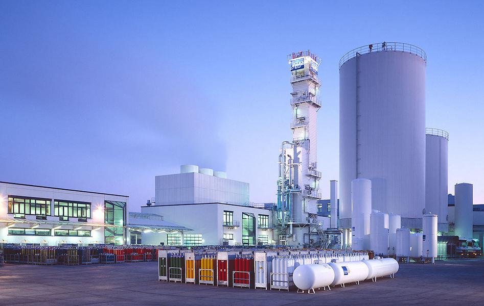 Ein Industriekomplex mit mehreren großen Behältern, Maschinen und Lagerräumen. Im Vordergrund stehen farbige Gaskontingente. Der Himmel ist hellblau, was auf Morgen- oder Abendlicht hinweist. Die Struktur zeigt einen modernen, funktionalen Baustil.