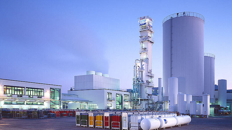 Ein Industriekomplex mit mehreren großen Behältern, Maschinen und Lagerräumen. Im Vordergrund stehen farbige Gaskontingente. Der Himmel ist hellblau, was auf Morgen- oder Abendlicht hinweist. Die Struktur zeigt einen modernen, funktionalen Baustil.