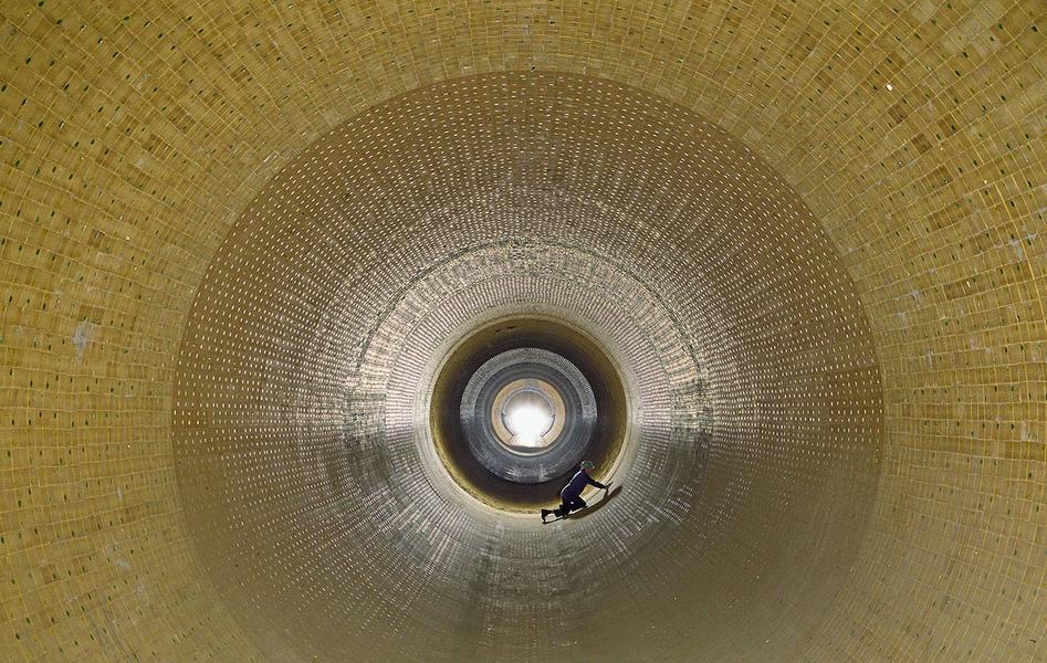 Eine perspektivische Ansicht des Innenraums eines großen, zylindrischen Behälters aus Steinen oder Platten. In der Mitte ist eine Lichtquelle sichtbar, während eine Person am Boden arbeitet. Die Wände sind rhythmisch strukturiert und reflektieren das künstliche Licht.