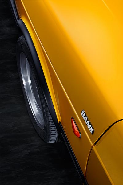 Ein gelber Saab-Auto mit einem glänzenden Lack. Die Aufnahme zeigt die Seitenansicht, wobei das Saab-Emblem und ein Rad sichtbar sind. Der Hintergrund ist dunkel und hebt die Farbe des Fahrzeugs hervor.