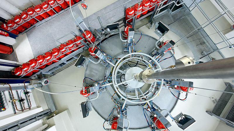 Eine Vogelperspektive auf eine kreisförmige Anordnung von technischen Geräten und Rohren. Um den zentralen Bereich sind mehrere rote Gasflaschen auf einer geraden Plattform angeordnet. Der Boden ist grau und die Umgebung wirkt industriell.