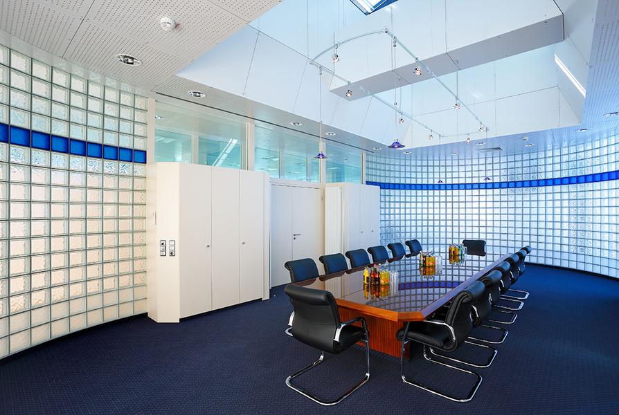 Ein moderner Besprechungsraum mit einem großen, rechteckigen Tisch in der Mitte, umgeben von schwarzen Stühlen. Die Wände sind mit transparenten Glasbausteinen dekoriert, und an der Decke befinden sich Fenster, die Tageslicht hereinlassen.
