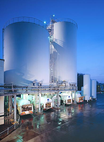 Zwei große weiße Silos sind bei Dämmerung zu sehen, umgeben von einer glatten, nassen Fläche. In der Nähe befinden sich mehrere Tanks, die in Betrieb sind, und LEDs leuchten in Blau. Die Atmosphäre wirkt industriell und modern.