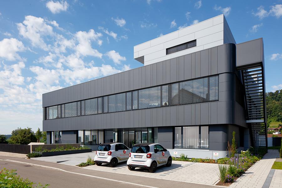 Modernes, mehrstöckiges Gebäude mit großen Fenstern und grauer Fassadenverkleidung. Vor dem Gebäude stehen zwei kleine Autos auf einem gepflasterten Parkplatz. Im Hintergrund sind Bäume und ein blauer Himmel mit einigen Wolken sichtbar.