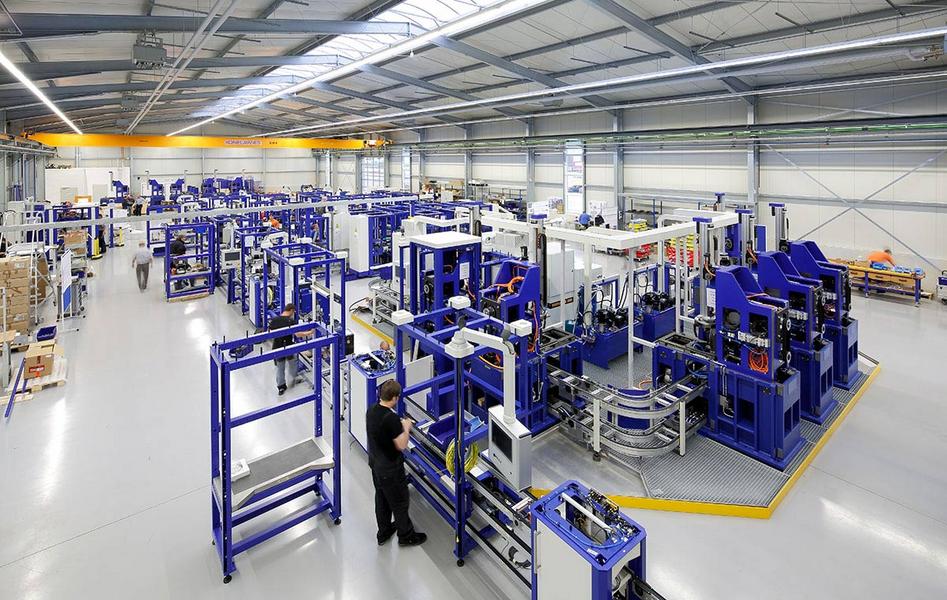 Eine weite Aufnahme einer modernen Produktionshalle, die mit verschiedenen Maschinen und Fertigungsstationen in Blau und Weiß ausgestattet ist. Arbeiter sind in der Halle aktiv. Der Raum hat hohe Decken und viel natürliches Licht, das durch große Fenster strömt.