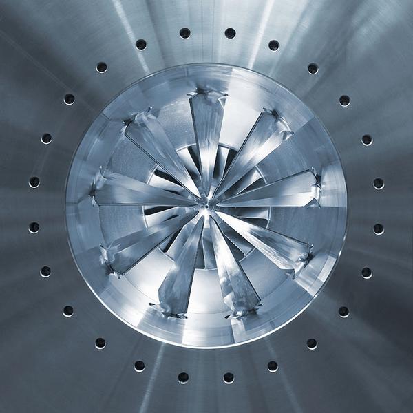 Ein runder metallischer Gegenstand mit einer spiralförmigen Anordnung von scharfen Klingen in der Mitte. Der Hintergrund ist silbern und glänzend, mit kleinen Löchern, die gleichmäßig um die Klingen herum angeordnet sind.