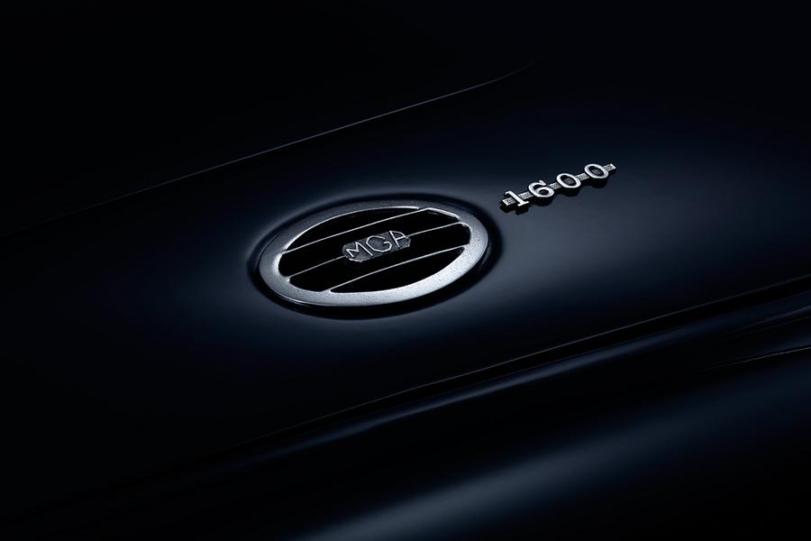 Ein schimmerndes Logo und die Modellbezeichnung "MGA 1600" auf einer dunklen Fahrzeugoberfläche. Das Logo hat eine runde Form mit horizontalen Streifen und ist in silberner Farbe gehalten.