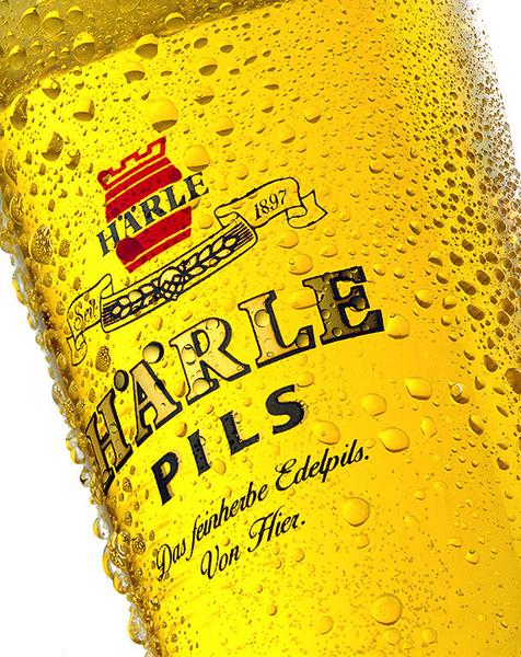 Ein Glas mit goldenem Bier, das reich an Blasen ist. Auf dem Glas steht der Schriftzug "HARLE PILS" und darunter der Slogan "Das feinherbe Edelpils. Von Hier." Hintergrund ist unscharf.