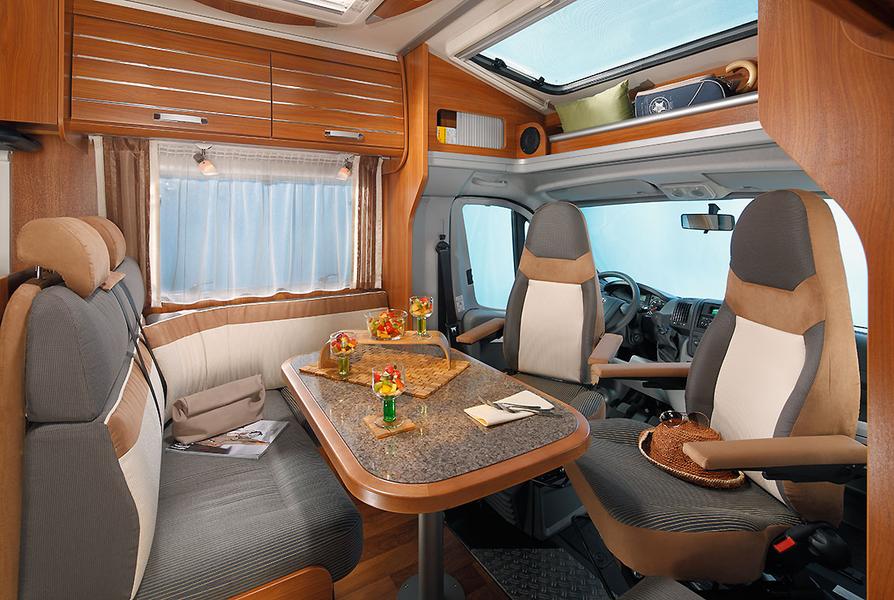 Innenansicht eines Wohnmobils mit einem Esstisch, umgeben von zwei Sitzgruppen. Auf dem Tisch stehen Snacks und Getränke. Die Wände sind mit Holzverkleidung ausgestattet, und große Fenster sorgen für genügend Licht. Im Hintergrund ist der Fahrersitz zu sehen.