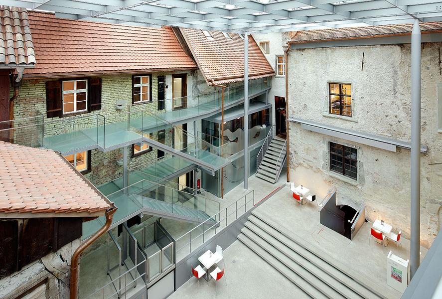 Ein Überblick über ein modernes Atrium mit transparenten Glasböden und einer Kombination aus historischen Gebäuden. Die Umgebung ist hell erleuchtet, mit Treppen und Sitzbereichen. Die Architektur verbindet Alt und Neu in harmonischer Weise.