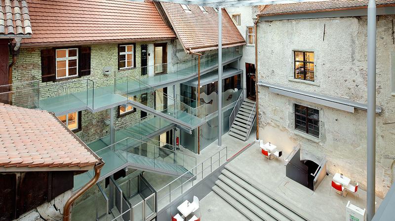 Ein Überblick über ein modernes Atrium mit transparenten Glasböden und einer Kombination aus historischen Gebäuden. Die Umgebung ist hell erleuchtet, mit Treppen und Sitzbereichen. Die Architektur verbindet Alt und Neu in harmonischer Weise.