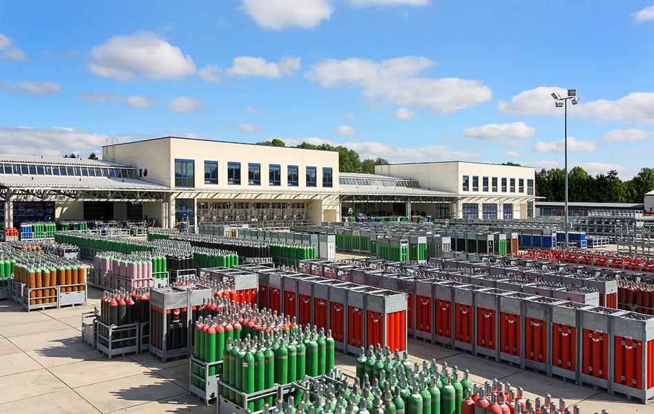 Eine weitläufige Lagerfläche mit zahlreichen Gasflaschen in unterschiedlichen Farben. Im Hintergrund sind moderne Gebäude und ein blauer Himmel mit wenigen Wolken zu sehen.