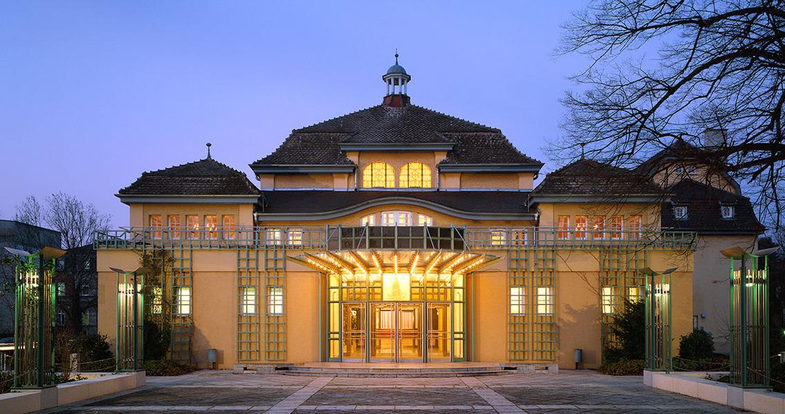 Historisches Gebäude mit stilvollem Eingang, beleuchtet in der Dämmerung. Die Fassade zeigt eine Mischung aus traditioneller und moderner Architektur, umgeben von Bäumen und einem geräumigen Vorplatz.