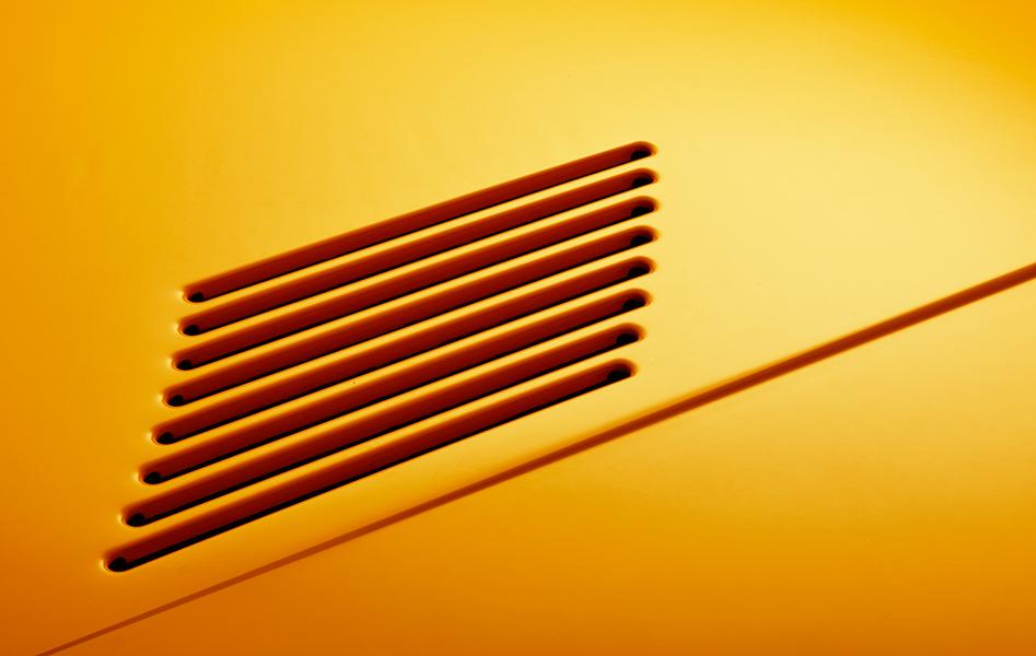 Detailaufnahme von einer orangefarbenen Oberfläche mit mehreren parallel angeordneten Schlitzöffnungen, die Schatten werfen.