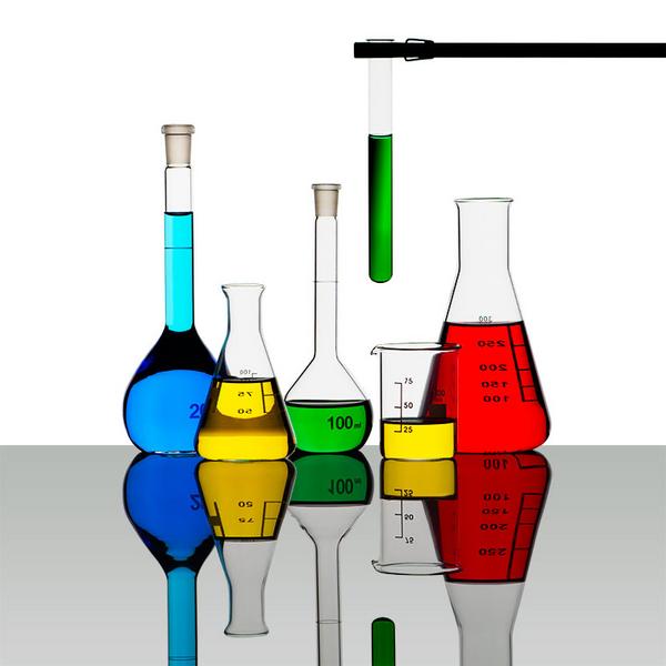 Eine Anordnung von fünf verschiedenen Chemikalien in Reagenzgläsern und Erlenmeyerkolben. Die Flüssigkeiten sind in verschiedenen Farben: Blau, Grün, Gelb und Rot. Die Gläser stehen auf einem reflektierenden Tisch.