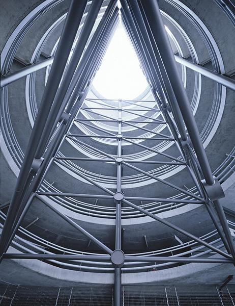 Ein Blick nach oben in einen modernen, zylindrischen Raum mit sichtbaren Stahlträgern. Das Licht strömt von oben durch eine kreisförmige Öffnung, während die Wände aus Beton und Stahl konstruiert sind. Die Struktur vermittelt ein Gefühl von Höhe und Raum.