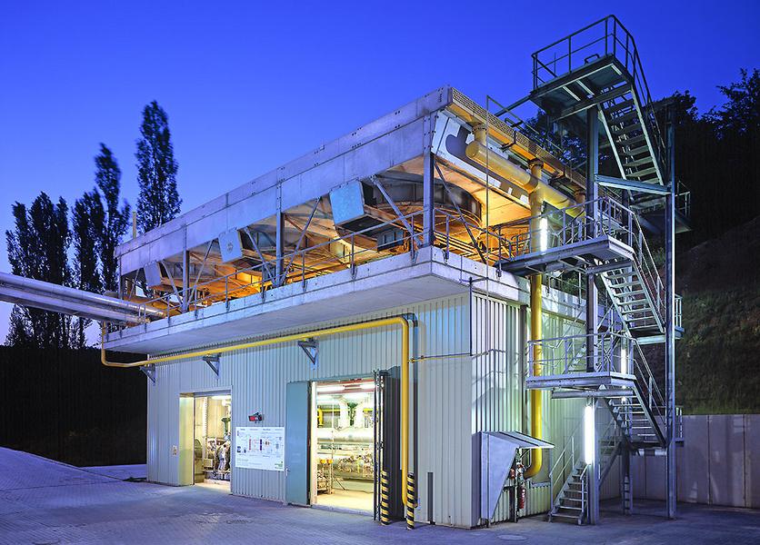 Eine moderne Industrieanlage bei Dämmerung, beleuchtet von gelbem und weißem Licht. Der Bau zeigt eine Stahlkonstruktion mit einer Treppe und mehreren Ebenen, umgeben von Bäumen im Hintergrund.