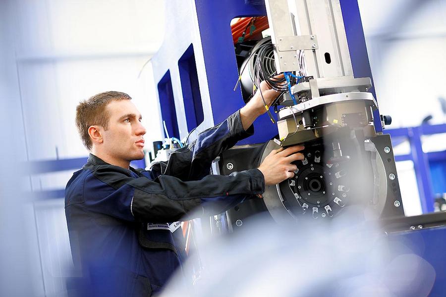 Ein Techniker arbeitet an einer Maschine in einer Industrieumgebung. Er hat eine konzentrierte Miene und hält eine Hand an einem Teil der Maschine, während er es anpasst. Die Umgebung ist modern und technisch gestaltet.