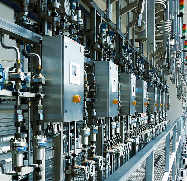 Nahaufnahme einer industriellen Anlagensteuerung mit mehreren silbernen Steuergeräten und Ventilen. Die Anordnung zeigt eine Reihe von Druck- und Temperaturmessgeräten sowie farbige Lichtanzeigen. Die Umgebung wirkt modern und technisch.