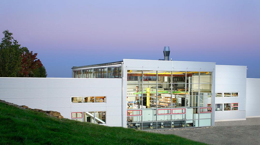 Moderne Architektur eines Gebäudes mit großen Glasfenstern. Die Fassade ist weiß, umgeben von Gras und Bäumen im Hintergrund. Der Himmel ist bei Dämmerung in sanften Farben gehalten.