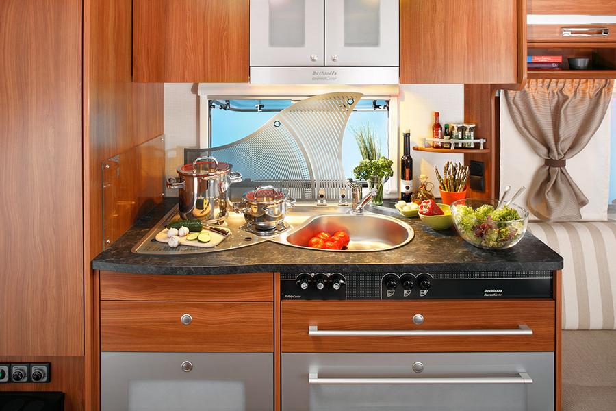 Küche in einem Wohnmobil mit Holzschränken. Ein runder Spülbecken ist in die Arbeitsplatte eingelassen, daneben stehen Töpfe und frisches Gemüse, darunter geschnittene Tomaten. Im Hintergrund sind Kräuter und Küchenutensilien sichtbar.