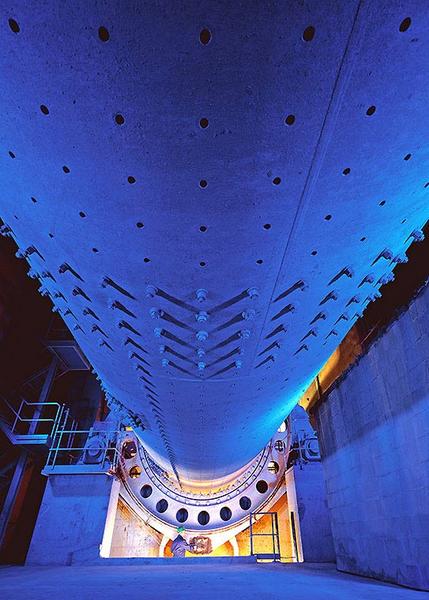 Blick auf die Unterseite eines Tunnelbohrers, beleuchtet in blauen und gelben Tönen. Die Struktur zeigt runde Löcher und mechanische Komponenten, die in einem unterirdischen Raum positioniert sind. Teile der Tunnelbau-Infrastruktur sind im Hintergrund sichtbar.