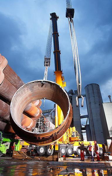 Ein Kran hebt ein großes Rohr an einer Baustelle in einer Industrieumgebung. Im Hintergrund sind weitere Baugeräte und ein bewölkter Himmel sichtbar. Arbeiter stehen in der Nähe und beobachten den Vorgang.