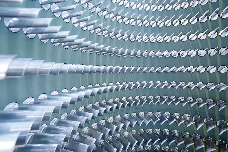 Eine abstrakte Nahaufnahme von glatten, metallischen Rohren, die in geschwungenen, parallel angeordneten Reihen angeordnet sind. Die Rohre reflektieren Licht und erzeugen eine dynamische, wellenartige Struktur. Der Hintergrund ist in sanften, gedeckten Farben gehalten.