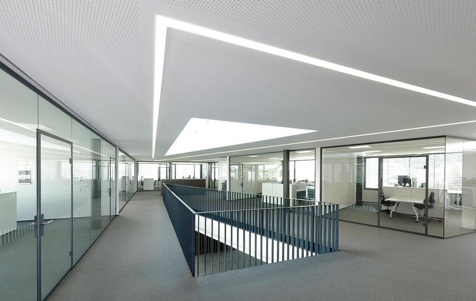 Moderne Bürolandschaft mit Glaswänden und einem offenen Bereich. Helle, minimalistische Gestaltung mit natürlichem Licht, das durch ein großes Dachfenster strömt. Ein erhöhter Gang führt zwischen den Büroräumen. Der Boden ist mit grauem Teppich ausgelegt.