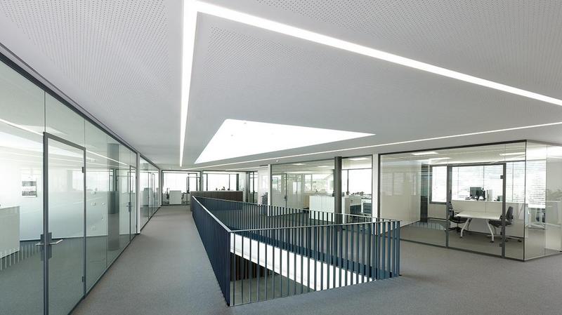 Moderne Bürolandschaft mit Glaswänden und einem offenen Bereich. Helle, minimalistische Gestaltung mit natürlichem Licht, das durch ein großes Dachfenster strömt. Ein erhöhter Gang führt zwischen den Büroräumen. Der Boden ist mit grauem Teppich ausgelegt.