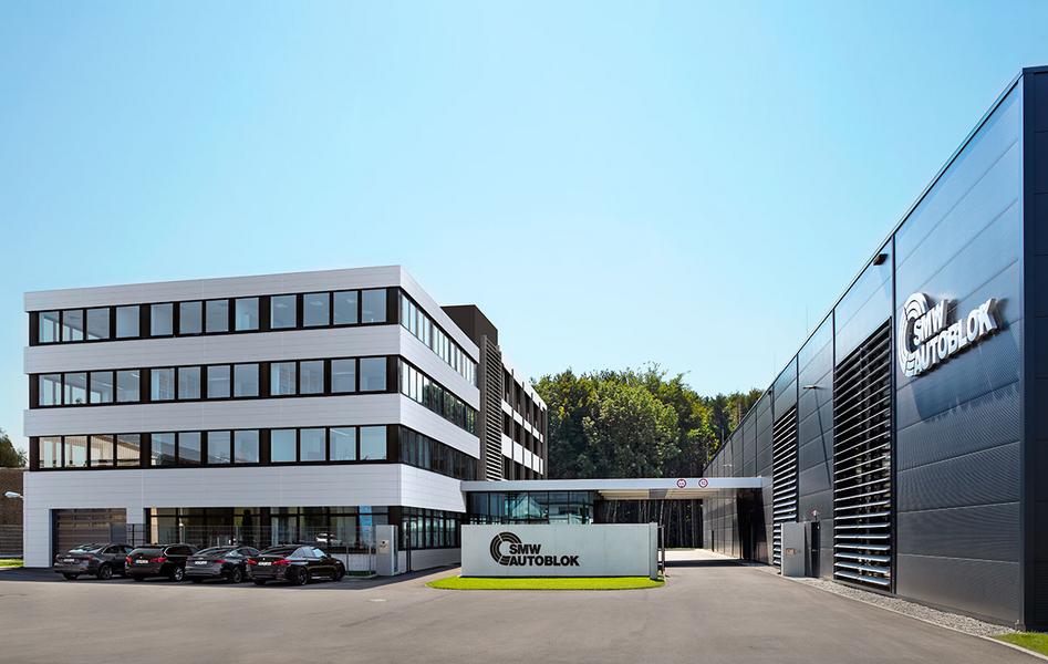 Moderne Unternehmensgebäude mit zwei Abschnitten, einem mehrstöckigen weißen Gebäude und einem schwarzen, modernen Anbau. Im Vordergrund steht ein Schild mit dem Firmenlogo. Vor dem Gebäude parken mehrere Autos. Der Himmel ist klar und blau.