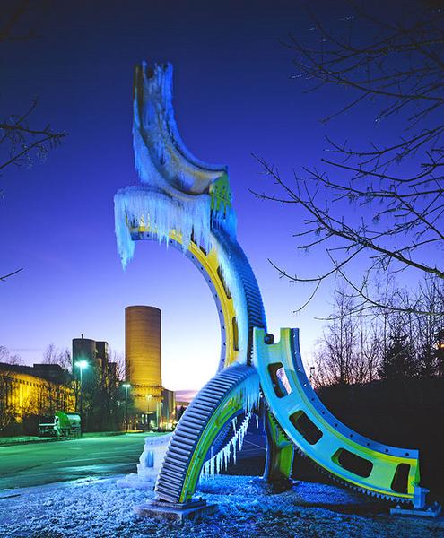 Eine große, bunte Skulptur in Form eines Zahnrads, das mit Eiszapfen bedeckt ist. Im Hintergrund sind Industriegebäude und ein Kühlturm sichtbar. Der Himmel ist in tiefen Blau- und Lilatönen erleuchtet, was eine kalte, aber beeindruckende Abendstimmung vermittelt.