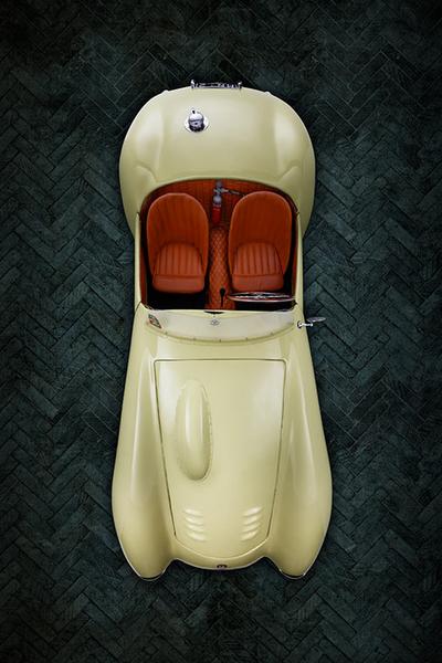 Ein cremefarbener Oldtimer mit roten Ledersitzen, aus der Vogelperspektive betrachtet. Der Wagen hat eine geschwungene Form und edle Details. Der Hintergrund ist aus dunklen, geflammten Platten gefertigt.