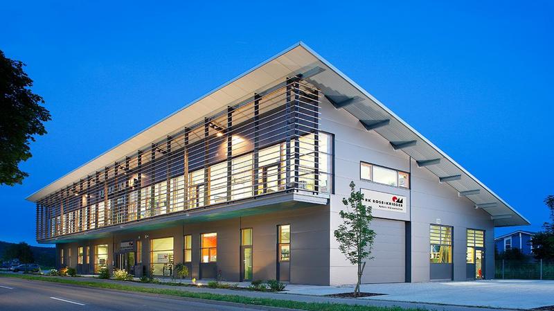 Moderner, zweigeschossiger Gebäude in der Dämmerung. Große Fenster und eine klare, rechteckige Struktur. Vor dem Gebäude ist ein kleiner Baum sowie eine asphaltierte Straße zu sehen. Die Fassade hat vertikale Holzleisten und eine klare Beschriftung am Eingang.