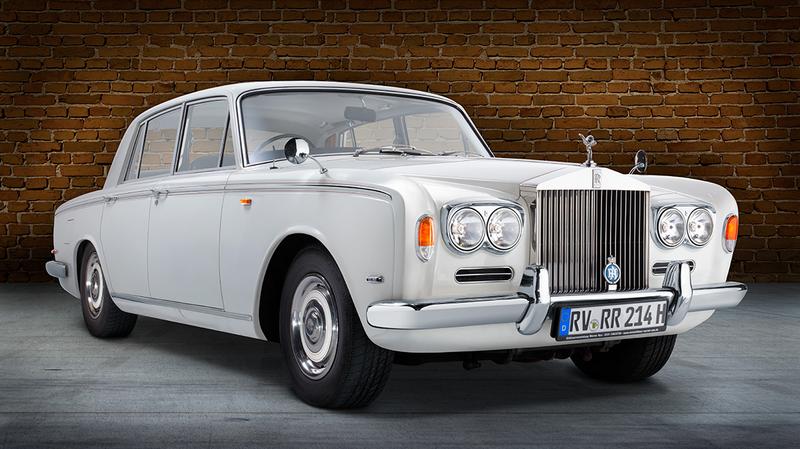 Ein klassisches weißes Auto, ein Rolls-Royce, steht vor einer Backsteinmauer. Die Front weist markante Scheinwerfer und das bekannte Kühleremblem auf. Das Fahrzeug hat eine elegante und luxuriöse Erscheinung.