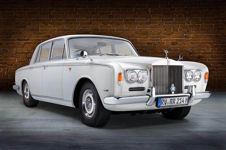 Ein klassisches weißes Auto, ein Rolls-Royce, steht vor einer Backsteinmauer. Die Front weist markante Scheinwerfer und das bekannte Kühleremblem auf. Das Fahrzeug hat eine elegante und luxuriöse Erscheinung.