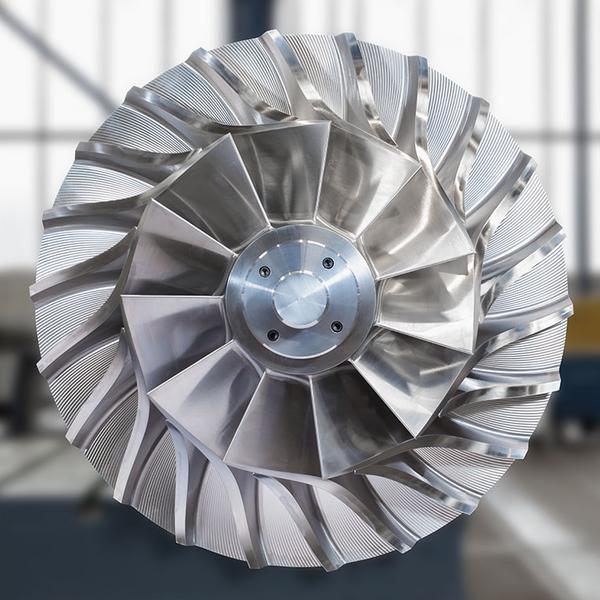 Detailansicht eines Turbinenrotors aus Metall. Die Rotorblätter sind symmetrisch angeordnet und haben eine geschwungene Form. Im Hintergrund sind unscharfe Werkstattelemente zu erkennen.