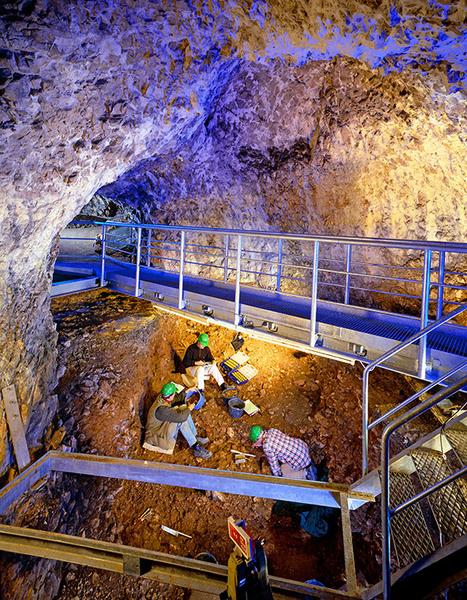 Eine Gruppe von Arbeitern mit grünen Helmen gräbt in einem Erdloch in einer Höhle. Über ihnen verläuft eine Metallbrücke, und die Wände der Höhle sind aus Stein. Die Beleuchtung erzeugt ein blaues Licht, das den Raum akzentuiert.