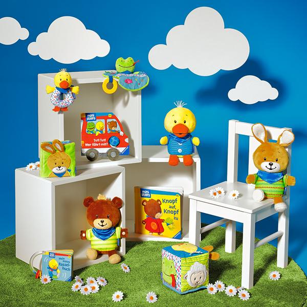 Bunte Spielzeuge und Plüschtiere auf einem grünen Teppich vor einer blauen Wand mit Wolken. Zu sehen sind ein Teddybär, eine Ente, ein Hase und weitere Spielartikel. Im Hintergrund stehen Bücher und ein Spielzeugauto.